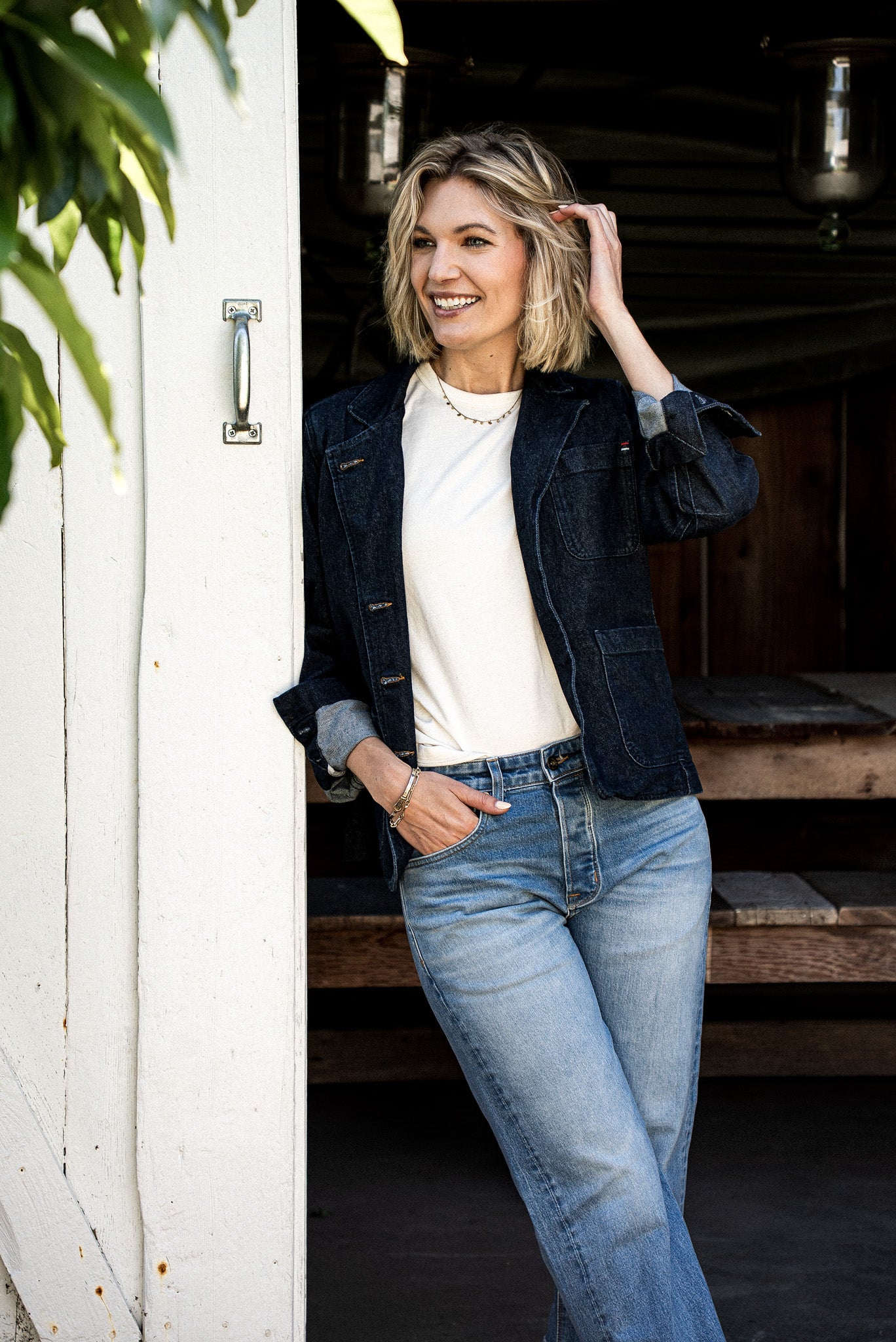 Woman in dark ocean-wash denim chore blazer over a cream tee and light jeans, leaning in a barn doorway.