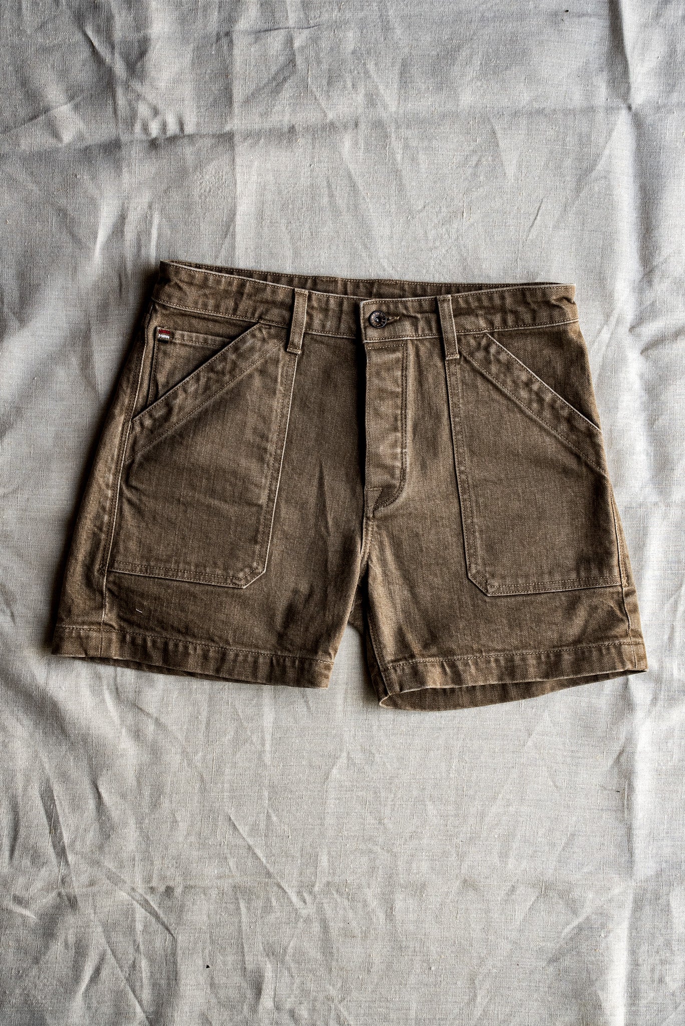 Wheat-toned Scout military denim shorts laid flat on natural linen, showing angled pockets.