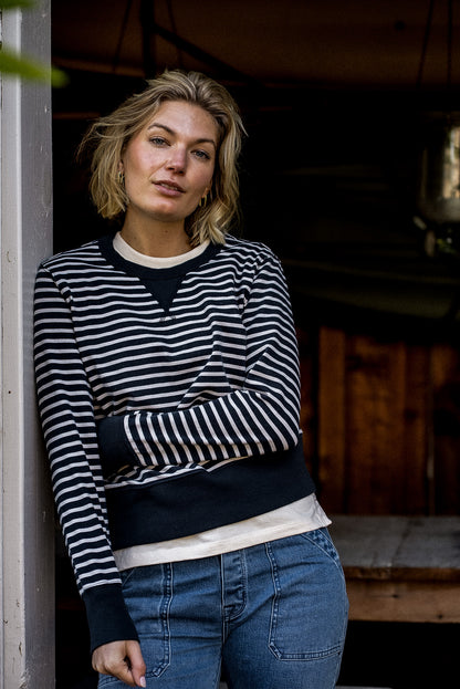 Woman in a black and cream striped Stinson rugby jersey pullover and blue jeans, leaning in a doorway.