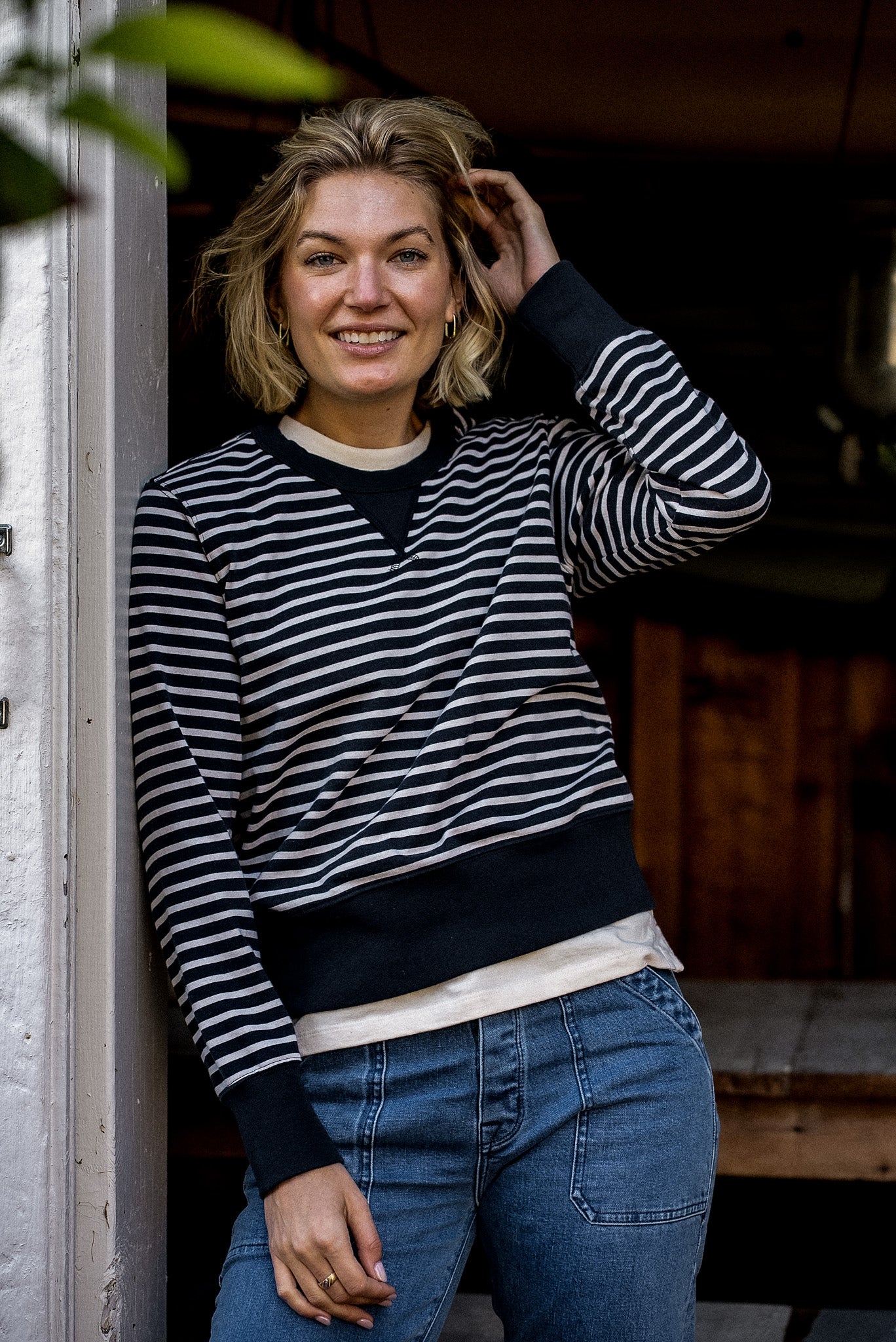 Smiling woman in a navy and cream striped Stinson rugby jersey pullover paired with blue jeans, leaning in a doorway.