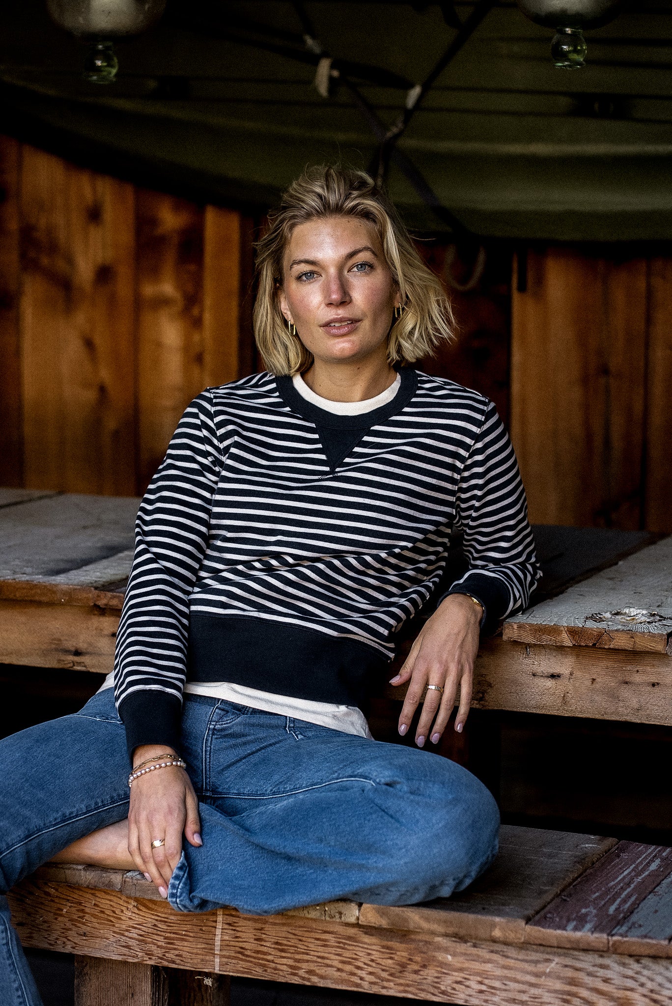 Woman in a black and white striped Stinson rugby jersey pullover and blue jeans seated on a wooden bench.
