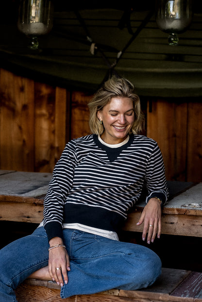 Woman in a black and white striped Stinson rugby jersey pullover and blue jeans, seated on a wooden bench.