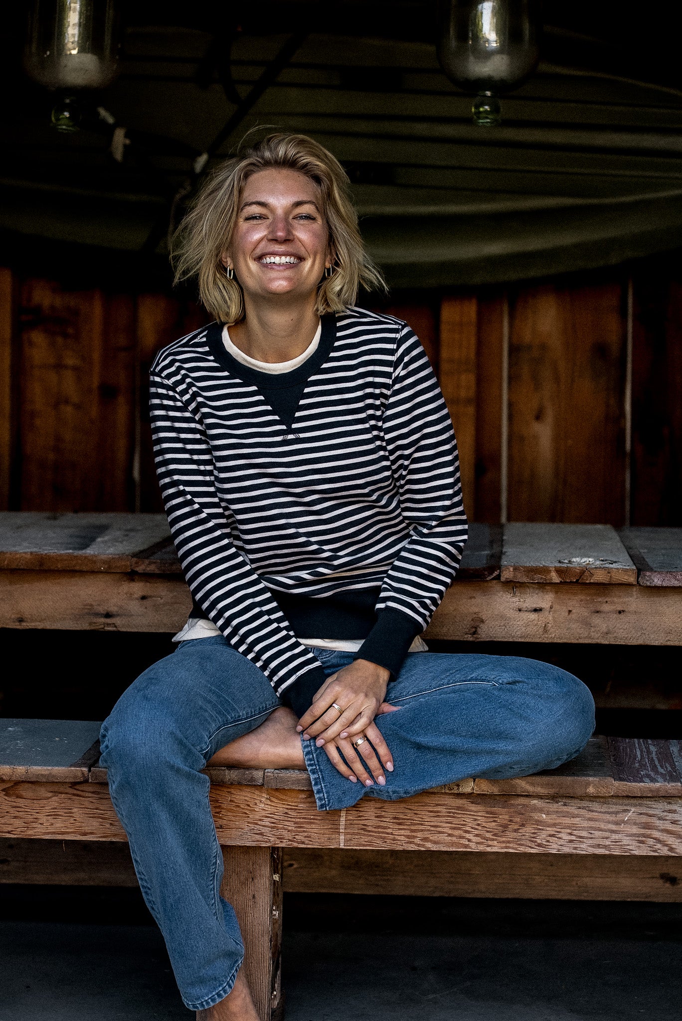 Woman smiling on a wooden bench wearing the navy and cream striped Stinson rugby jersey pullover with blue jeans.