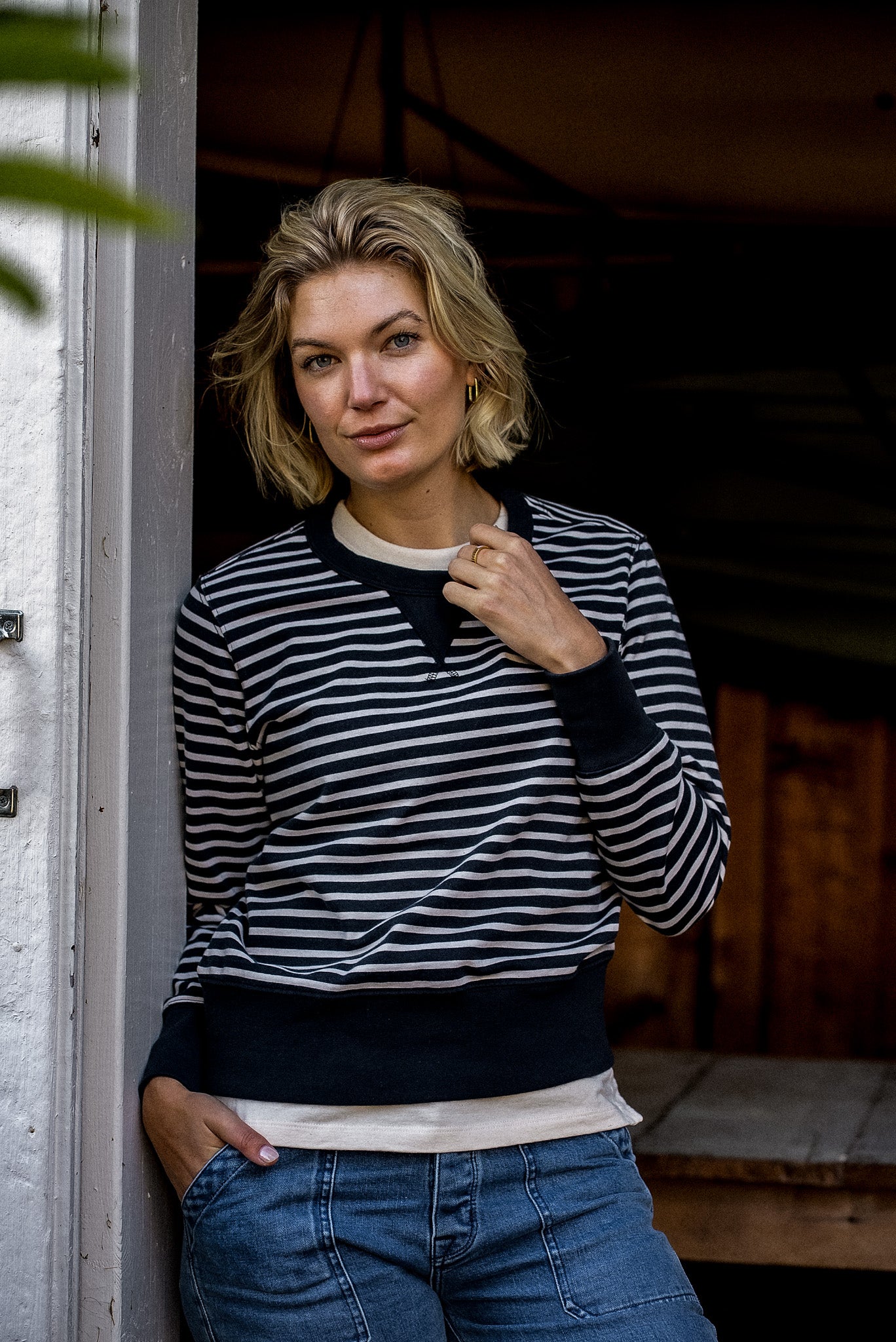 Woman in a black and cream striped Stinson rugby jersey pullover and denim jeans, leaning in a doorway.