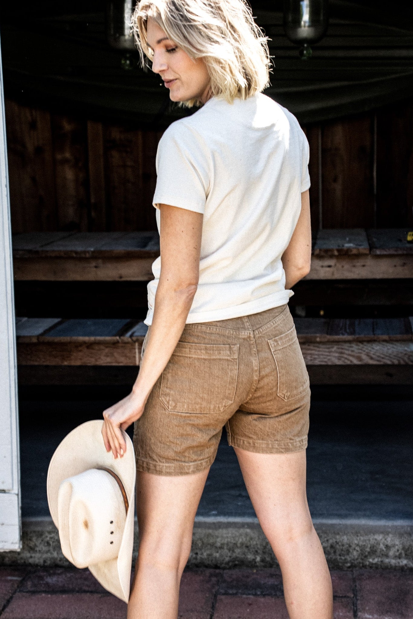 Back view of wheat denim military shorts with cream tee, holding a straw hat outdoors.