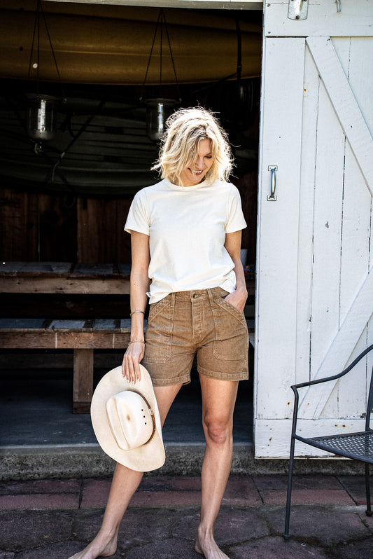 Woman in wheat-colored Scout denim military shorts and cream tee, holding a straw cowboy hat by a barn door.
