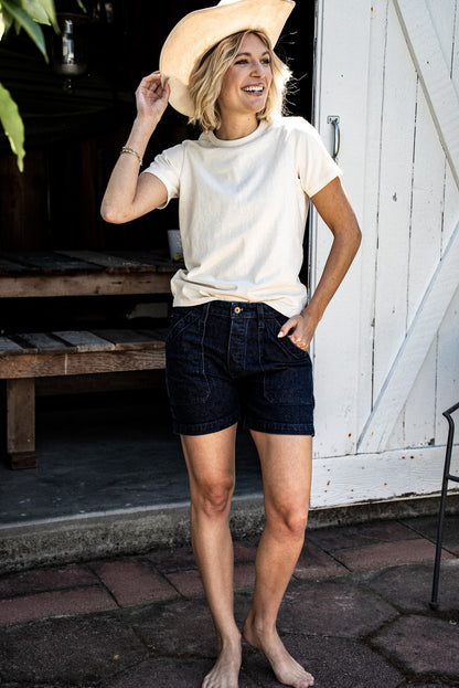 Woman in a cream tee and straw cowboy hat wearing Scout ocean-wash denim military shorts outside a barn door.