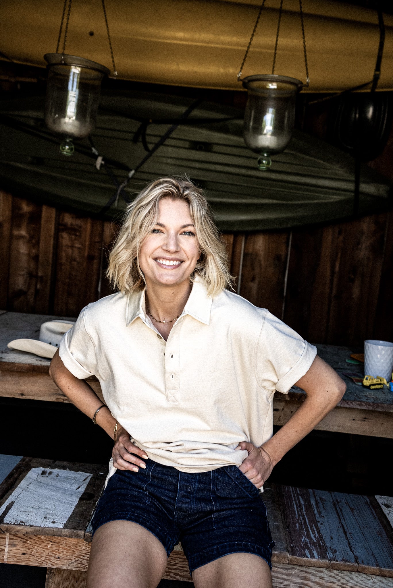 Smiling woman wearing Scout ocean-wash denim shorts and a cream polo, leaning against a rustic wooden table.