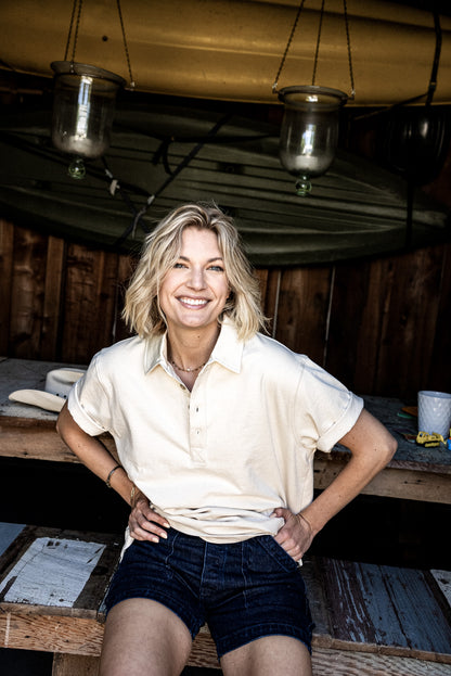 Smiling woman wearing Scout ocean-wash denim shorts and a cream polo, leaning against a rustic wooden table.