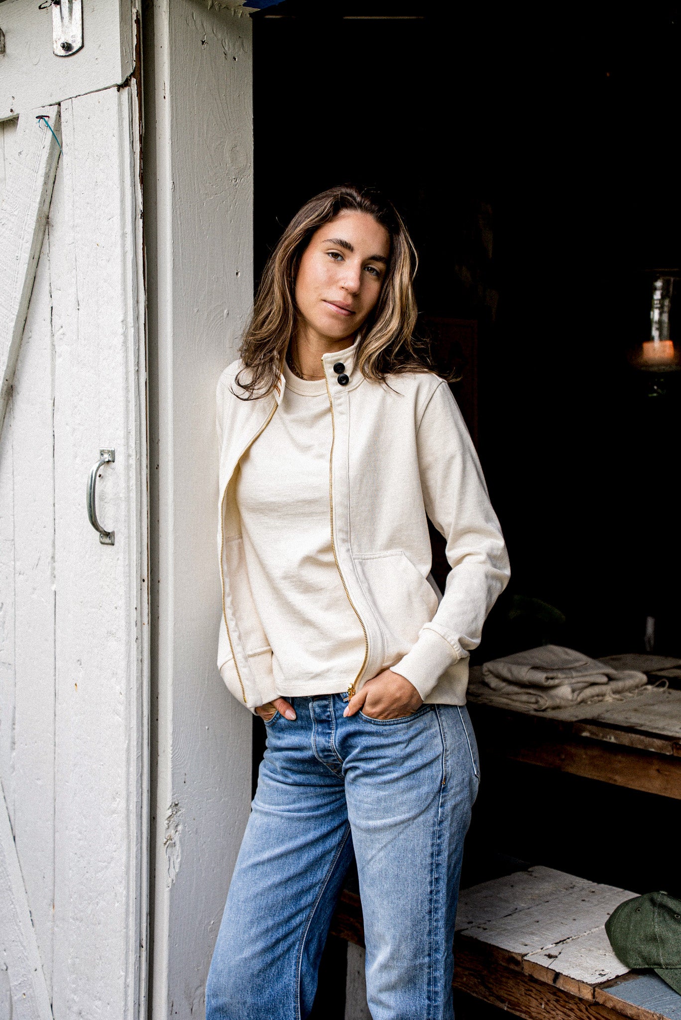 Person in a natural cotton moto jacket with asymmetric brass zipper, leaning in a doorway.