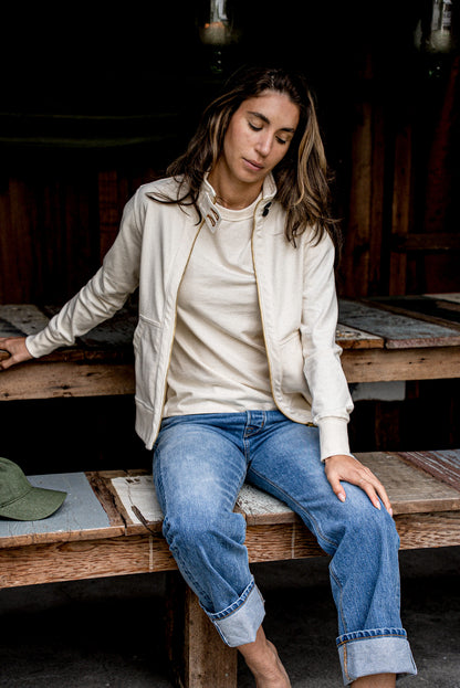 Natural cotton moto jacket worn open with brass zipper over blue jeans on a bench.