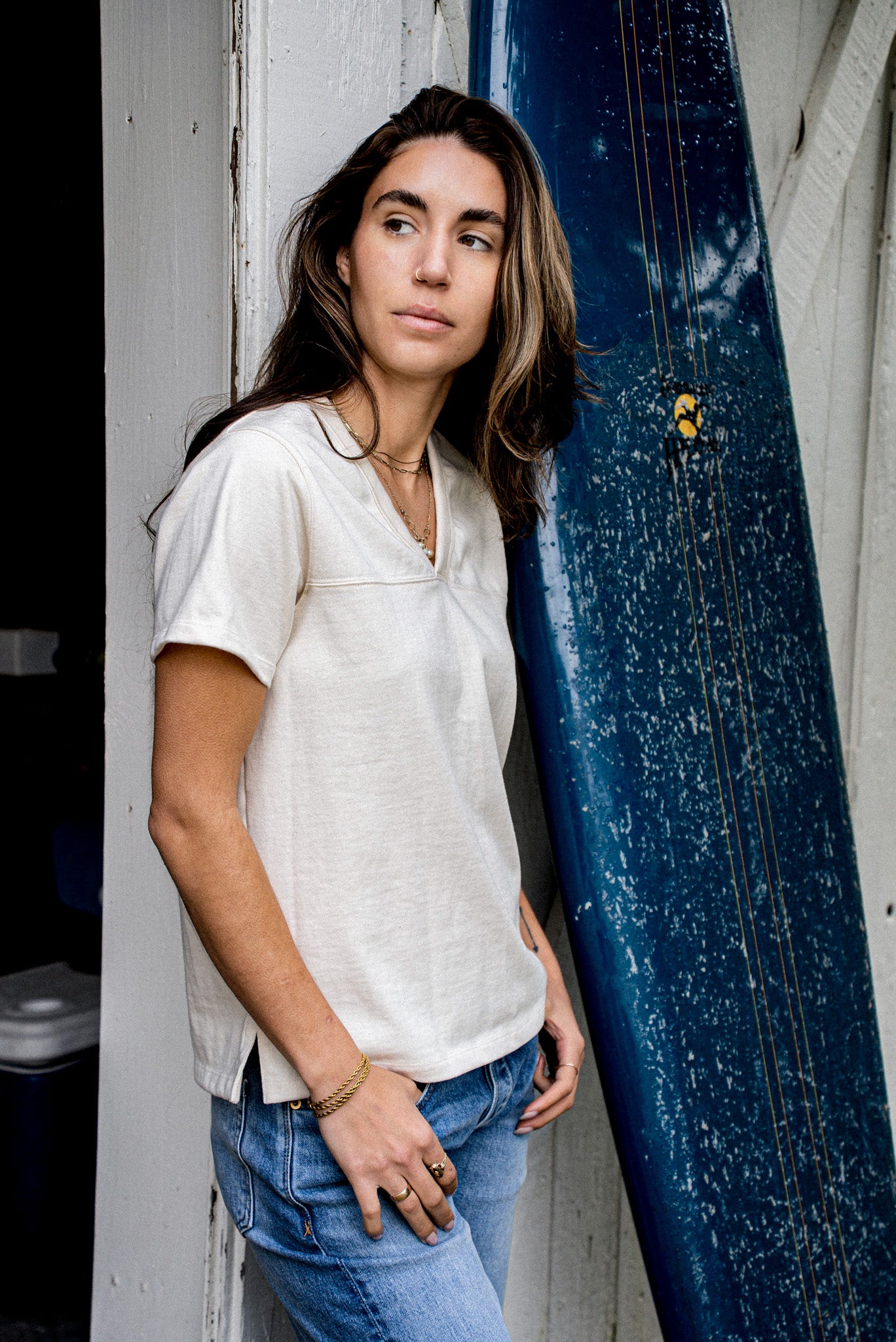 Model wears natural cotton Stinson V-neck jersey tee leaning beside a blue surfboard.