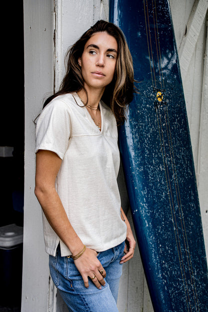 Model wears natural cotton Stinson V-neck jersey tee leaning beside a blue surfboard.