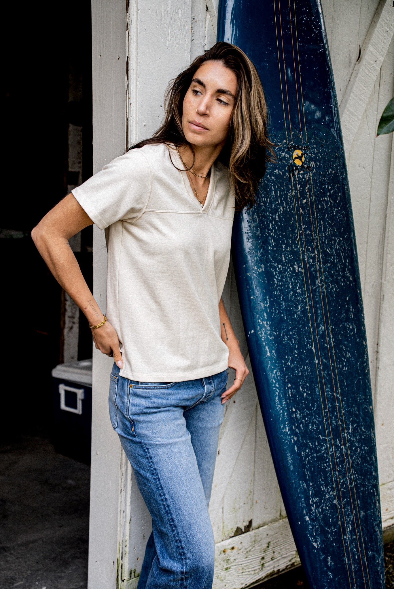 Model in natural cotton Stinson V-neck jersey leaning beside a blue surfboard.