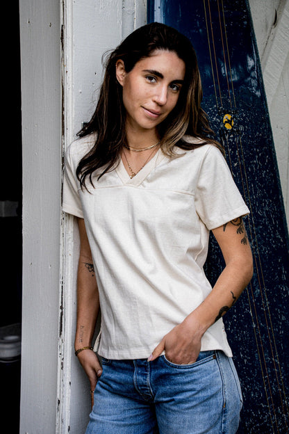 Woman in a natural cotton V-neck jersey tee, leaning against a weathered doorway.