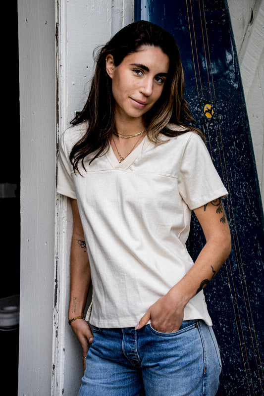 Woman in a natural cotton V-neck jersey tee, leaning against a weathered doorway.