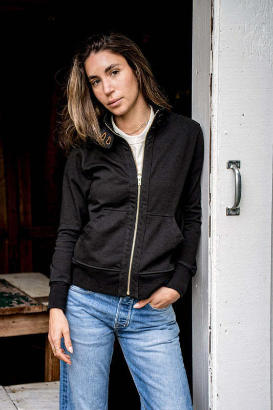 Woman in a black cotton moto jacket with brass zipper, leaning in a doorway.