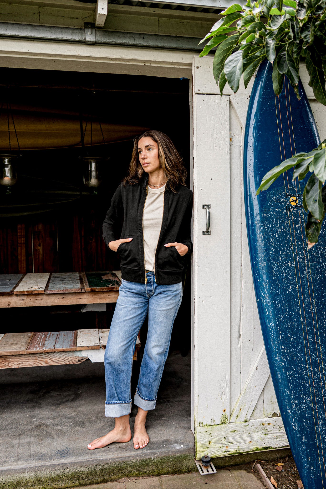 Woman wearing a black Stinson moto cotton jacket, zipped, standing by a surfboard.