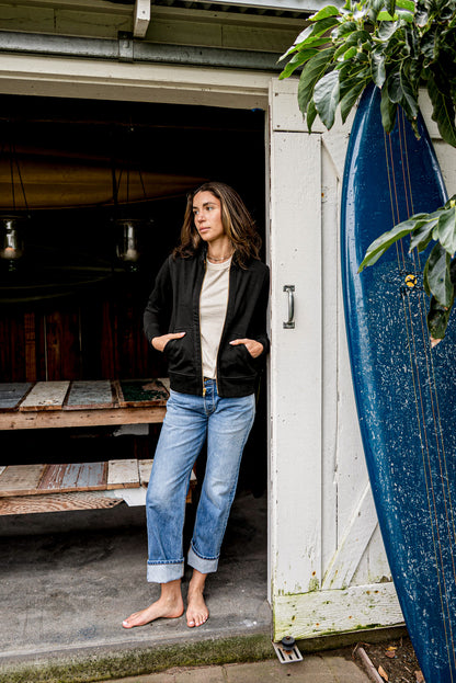 Woman wearing a black Stinson moto cotton jacket, zipped, standing by a surfboard.