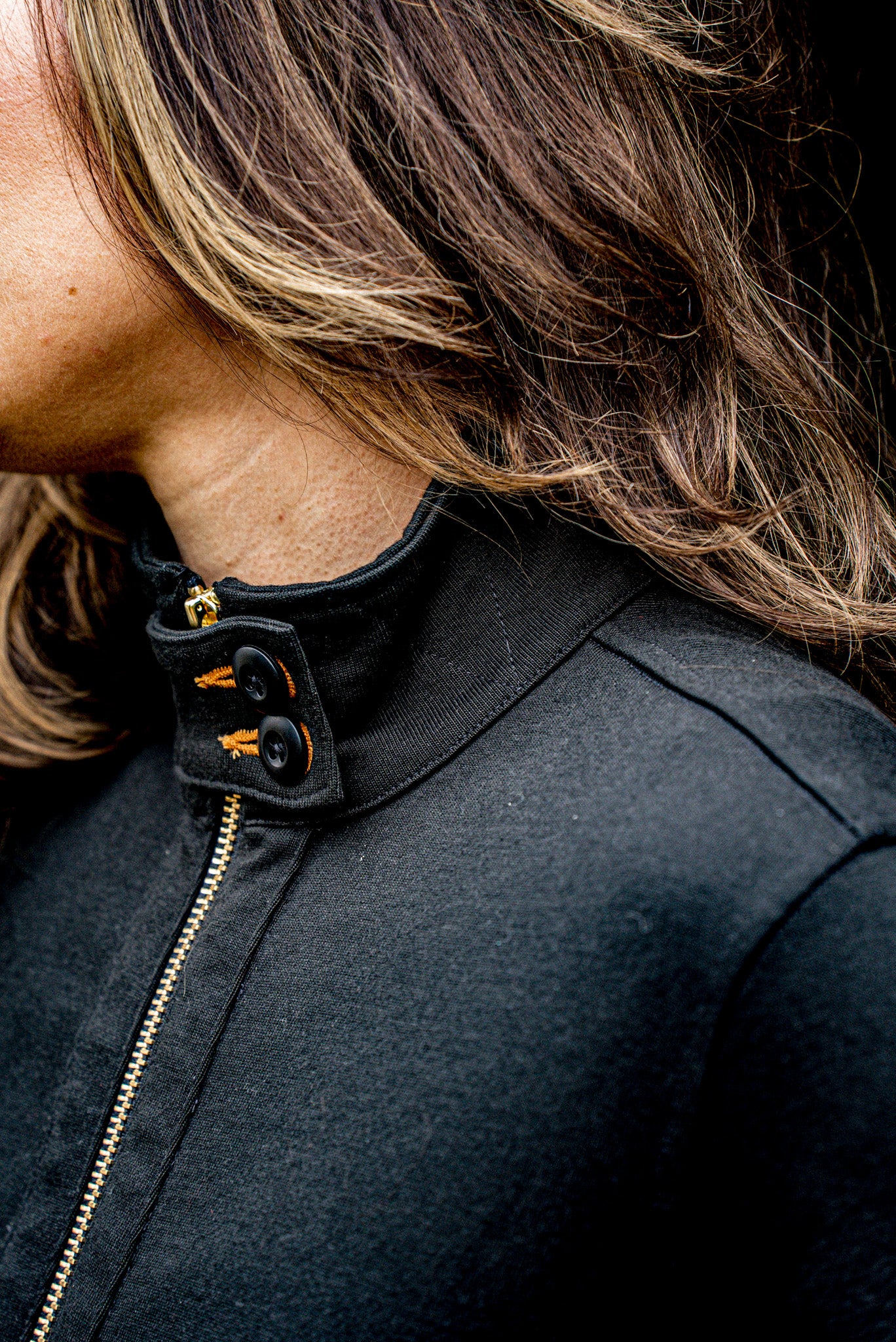 Close-up of black cotton moto jacket collar with brass zipper and buttoned tab detail.
