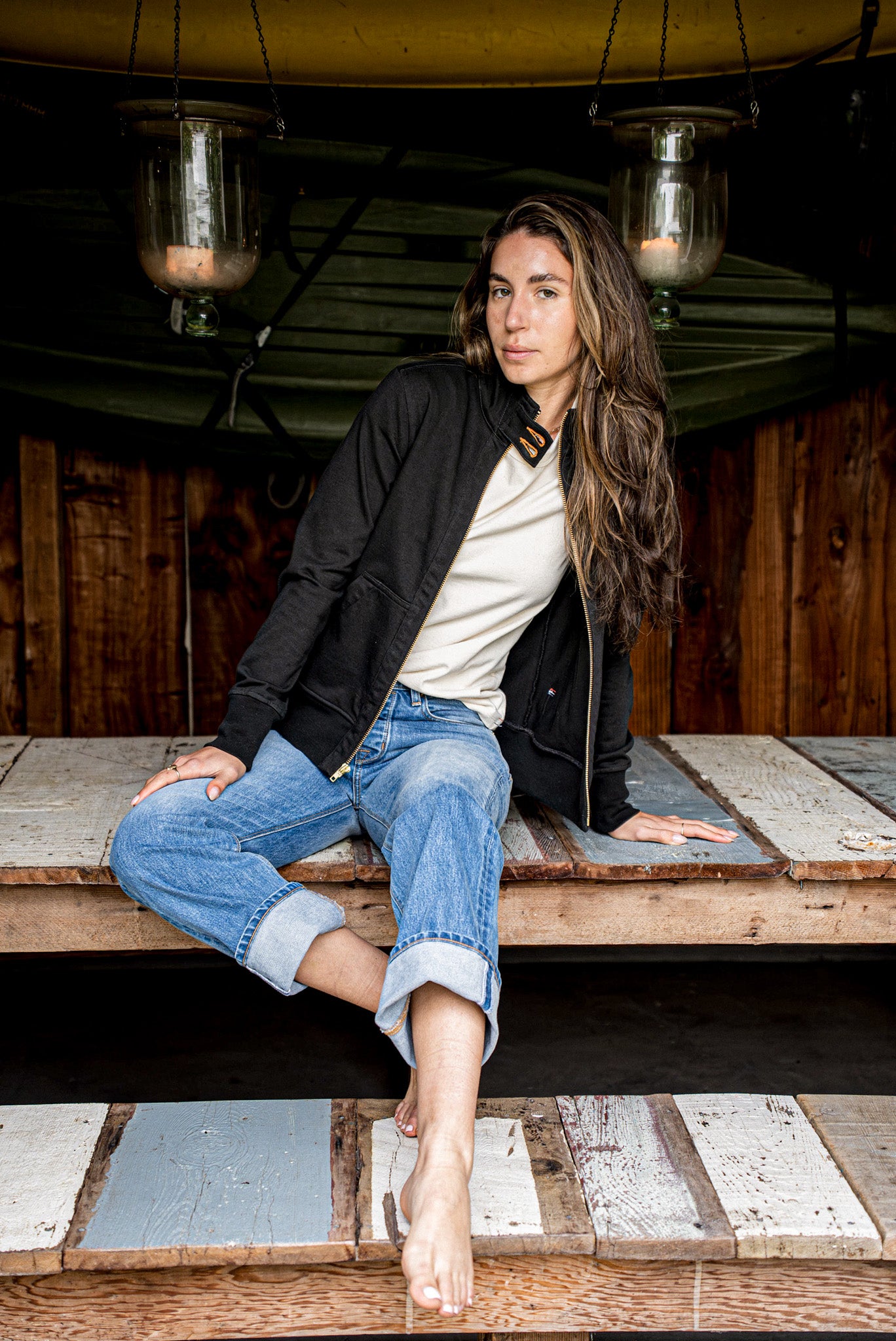Black Stinson moto jacket worn open over a cream tee, seated on weathered wood planks.