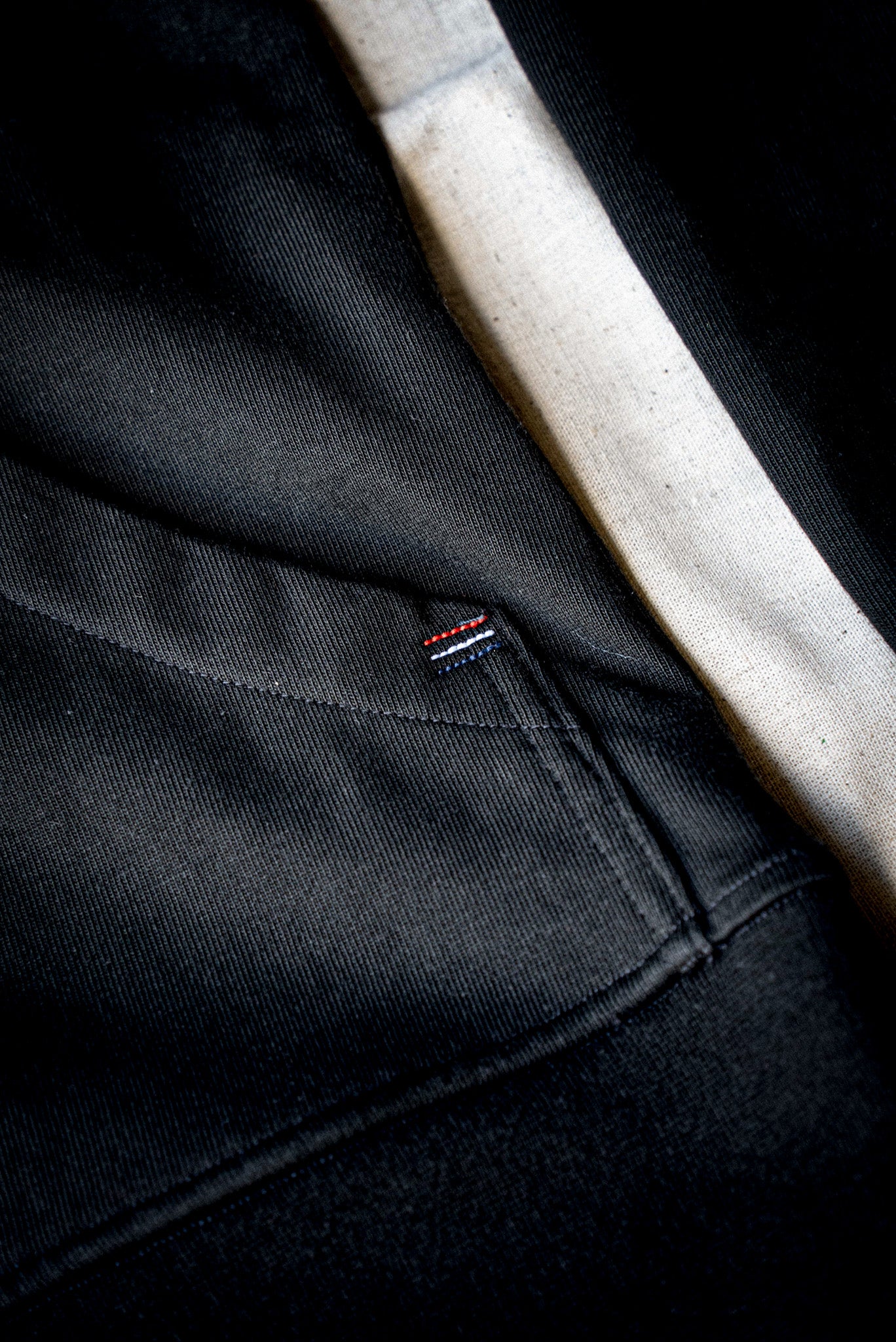 Close-up of black cotton moto jacket showing ribbed hem, seam stitching, and small tricolor tab.
