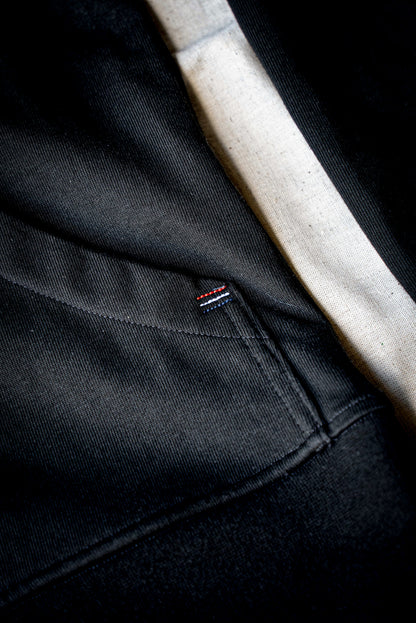 Close-up of black cotton moto jacket showing ribbed hem, seam stitching, and small tricolor tab.