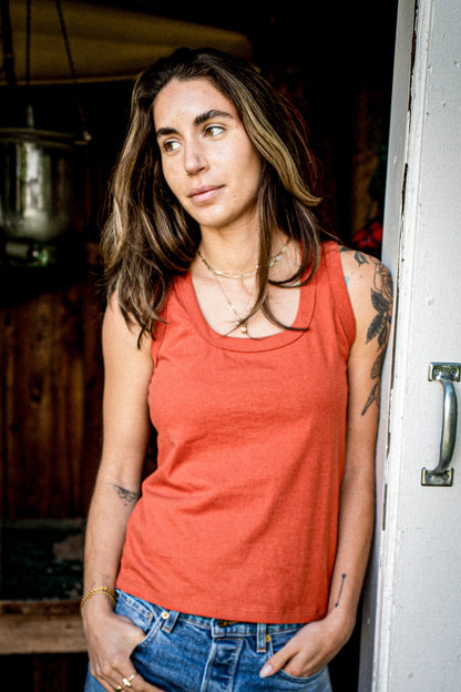 Woman in a brick red cotton Reyes tank and blue jeans leaning in a weathered doorway.