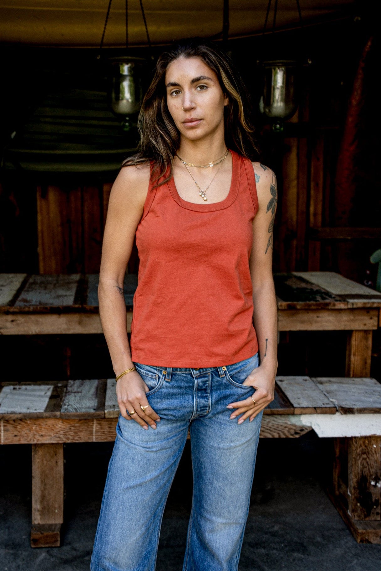Model in a brick-red cotton Reyes tank and faded blue jeans, standing by a wooden workbench.