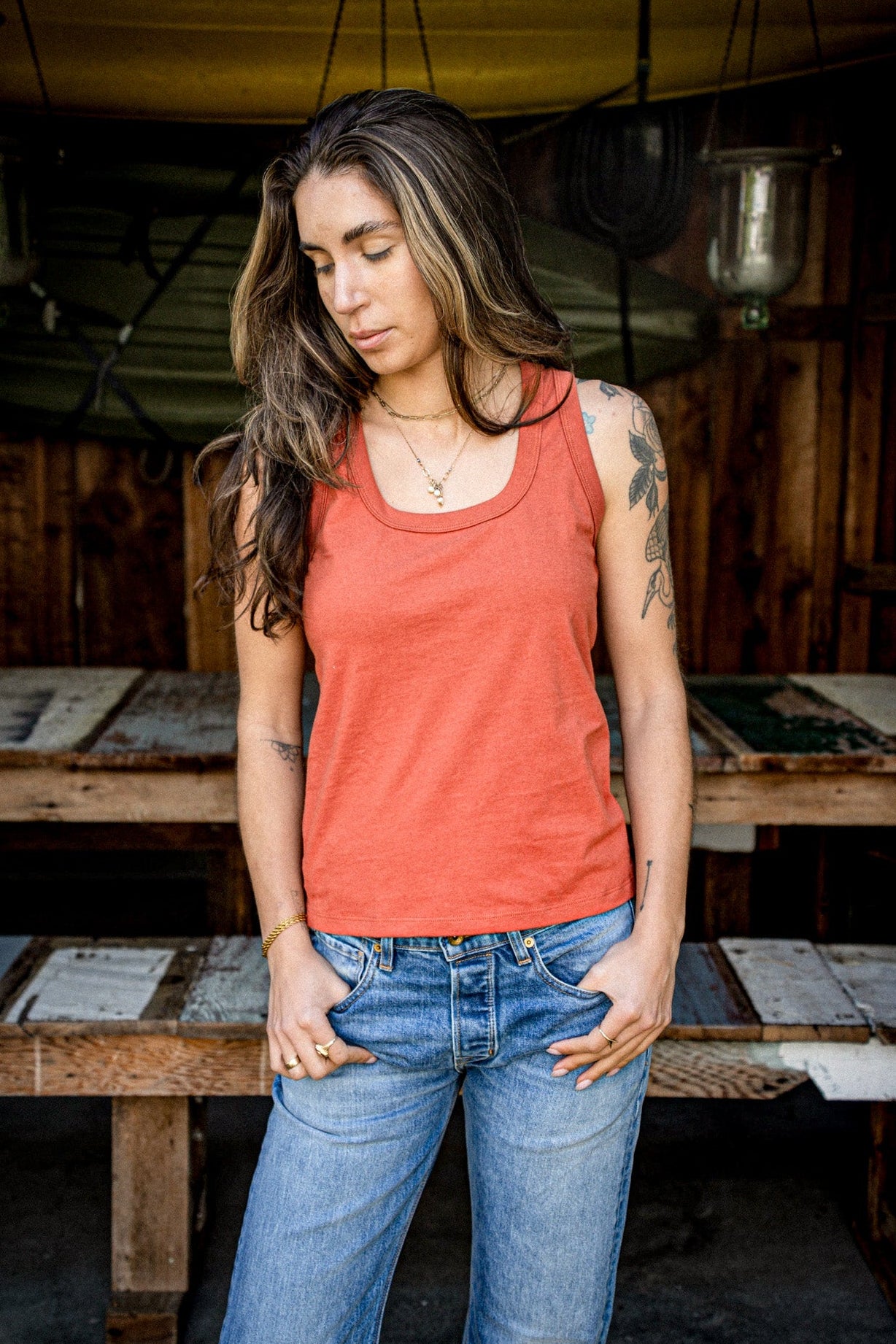 Woman wearing a brick-red scoop-neck cotton tank top with faded blue jeans in a rustic wooden workshop.
