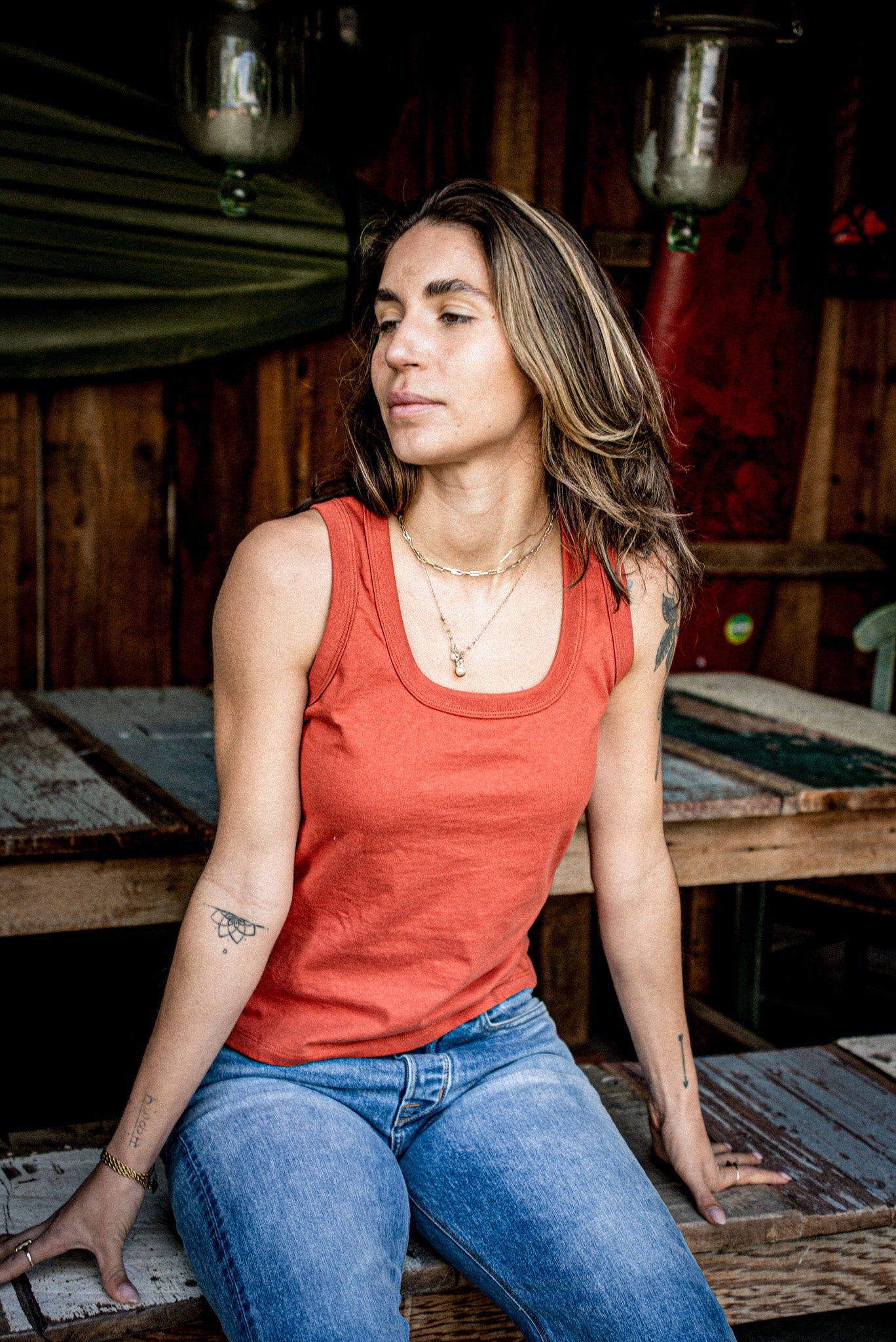 Woman in a brick-red Reyes cotton tank top and blue jeans seated on a weathered wooden bench.