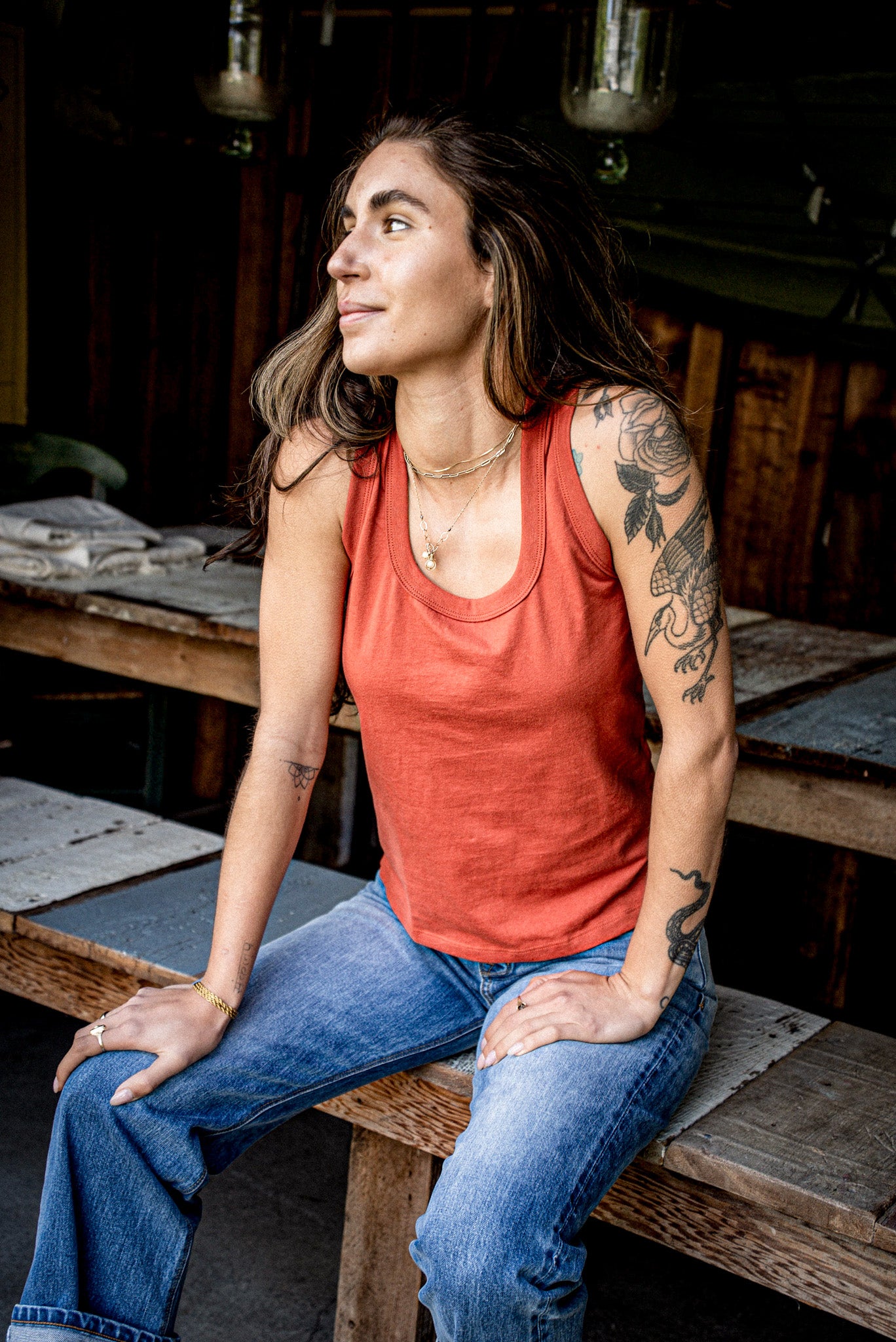 Woman in a brick red cotton Reyes tank top and blue jeans seated on a wooden workbench.