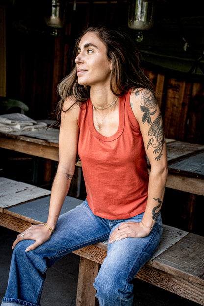 Woman in a brick red cotton Reyes tank top and blue jeans seated on a wooden workbench.