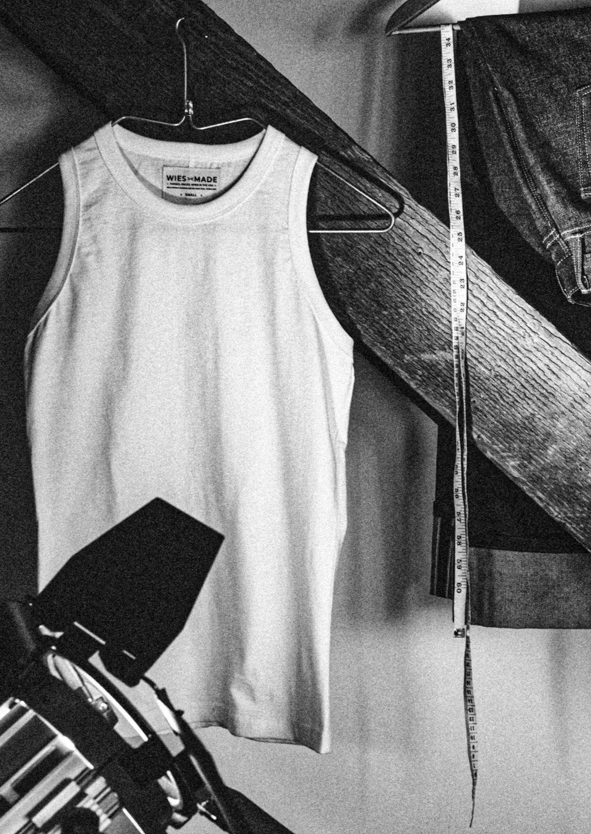 Bolinas cotton tank top on a hanger beside selvedge denim jeans and a measuring tape in a black-and-white studio scene.