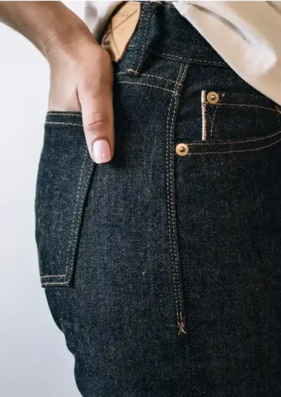 sidney Fit Jean in Raw Selvedge Denim color, featuring high-rise and straight-leg styling, worn by a woman with a tucked-in white shirt. The focus is on the back pocket and stitching details against a clean, neutral white background