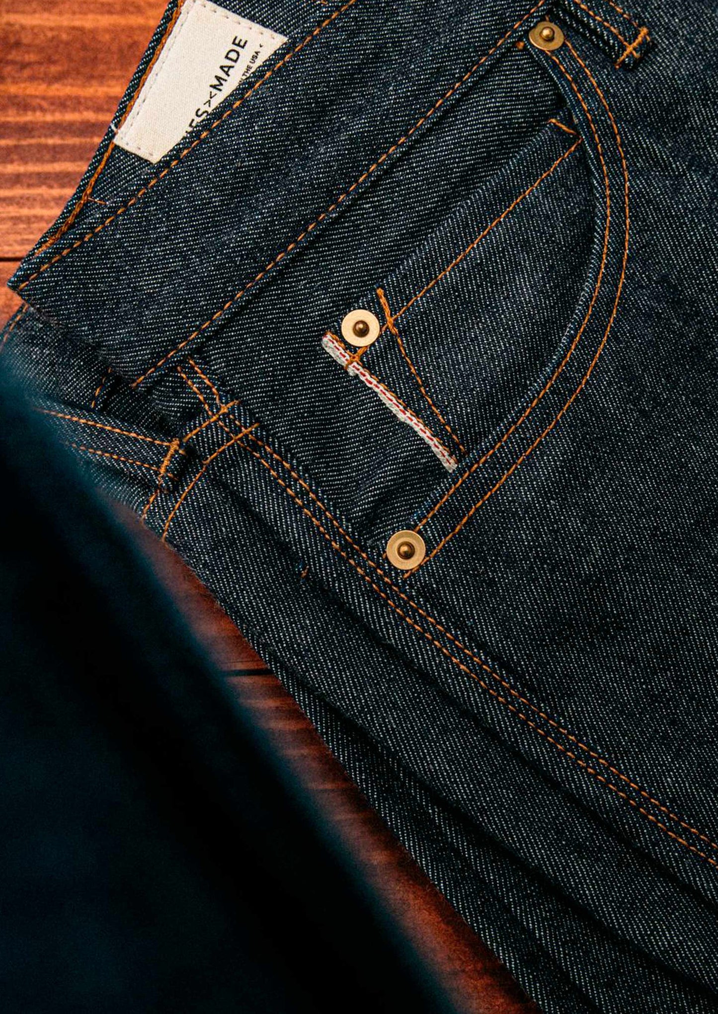 Men’s raw selvedge denim jeans shown in close-up, featuring dark indigo fabric with orange contrast stitching, brass rivets, and red-edge detailing that highlights authentic craftsmanship