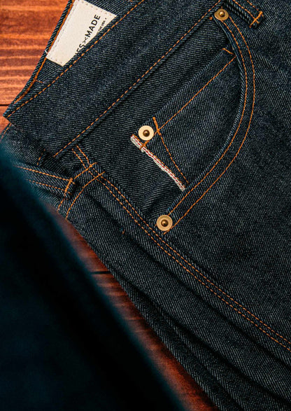 Men’s raw selvedge denim jeans shown in close-up, featuring dark indigo fabric with orange contrast stitching, brass rivets, and red-edge detailing that highlights authentic craftsmanship