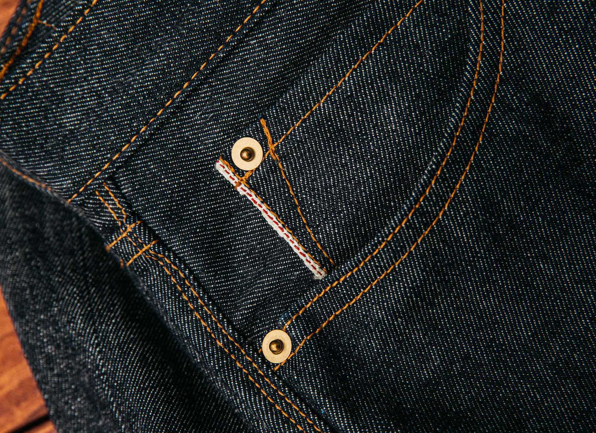 Selvedge denim jeans for men shown in a close-up, featuring tightly woven dark indigo fabric, brass rivets, and signature red-edge detailing that highlights craftsmanship and durability