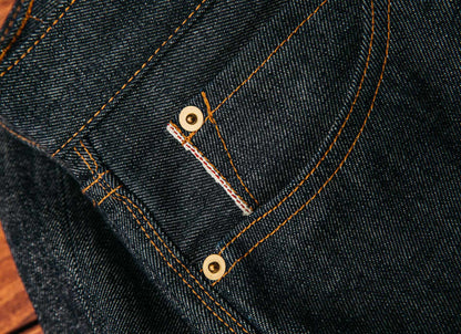 Selvedge denim jeans for men shown in a close-up, featuring tightly woven dark indigo fabric, brass rivets, and signature red-edge detailing that highlights craftsmanship and durability
